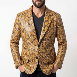 May include: A blazer featuring a yellow and brown snakeskin pattern with a notched lapel, two black buttons at the front closure, and two flap pockets at the waist. The garment is worn over a dark grey v-neck shirt and dark denim jeans.