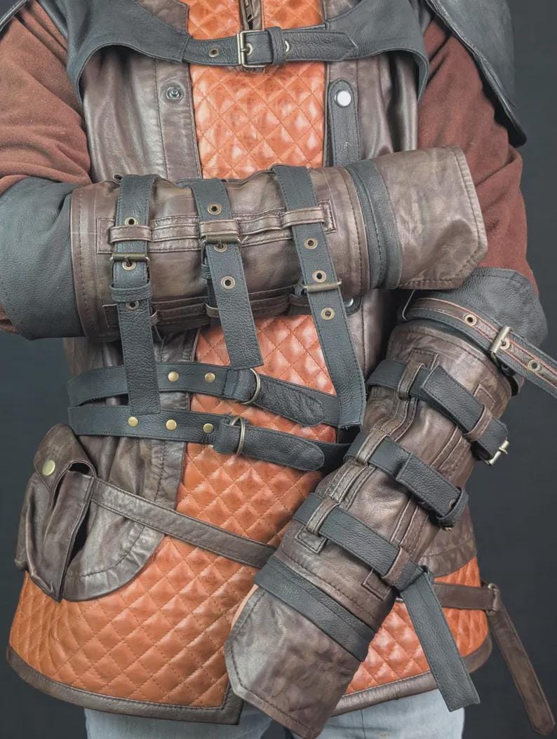 Op de afbeelding: Een bruin en zwart lederen vest met een gewatteerd oranje middenpaneel, meerdere gespriemen en ge&iuml;ntegreerde armbeschermers. Het kledingstuk is voorzien van donkere lederen overlays, metalen gespen en klinknagels, met een kleine zijzak.