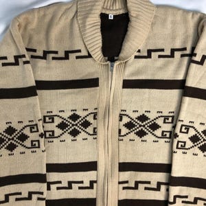 May include: A beige knit zip-up cardigan featuring a dark brown geometric pattern across the chest and sleeves. The garment has a ribbed shawl collar, a full-length front zipper, and a white interior tag labeled XL near the neckline.