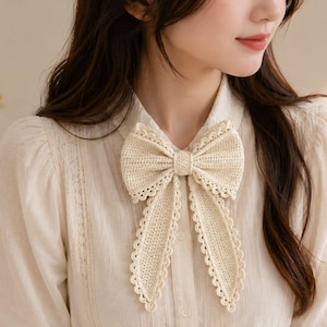 Può includere: Un fiocco all'uncinetto color crema caratterizzato da un motivo a maglia testurizzato e un delicato bordo in pizzo smerlato. L'accessorio è pensato per essere indossato sul colletto di una camicia, con un nodo centrale e due code lunghe.