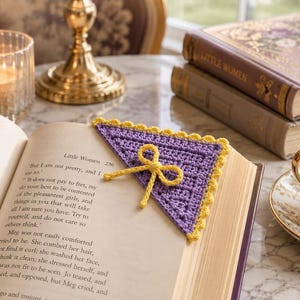 May include: A triangular crochet corner bookmark in lavender with a yellow scalloped border and a small yellow bow attached to the center, placed on the open page of a book titled Little Women, featuring visible printed text on the page.