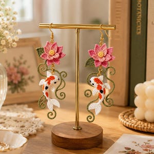 May include: A pair of dangling earrings featuring crocheted pink lotus flowers with green leaves and white, orange, and black koi fish motifs. The earrings are suspended from gold-toned metal hooks and hang from a gold-toned T-shaped display stand.