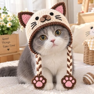 May include: A cream-colored crochet cat hat featuring dark brown ear tips with pink centers, embroidered black eyes, a nose, and whiskers. The hat includes two long, braided cream and brown earflaps ending in pink and brown crochet paw pads.
