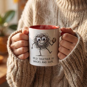 May include: A white ceramic mug with a red interior featuring a black line drawing of a scribbled character with googly eyes holding a chocolate chip cookie. The text below the character reads, "HELD TOGETHER BY SNACKS AND HOPE."