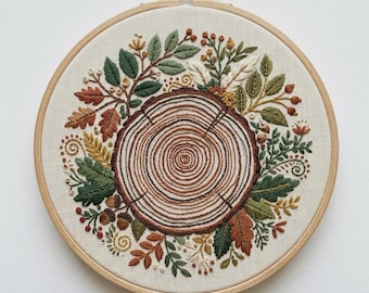 May include: A circular embroidery hoop containing a fabric piece with a hand-stitched design of a brown tree stump cross-section surrounded by a wreath of green leaves, orange oak leaves, small red berries, and brown acorns on a cream background.