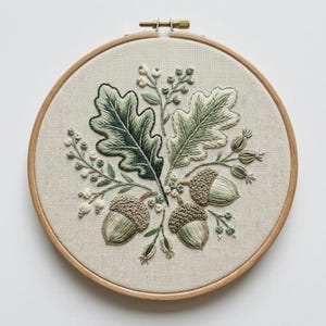 May include: A circular wooden embroidery hoop framing a piece of off-white fabric featuring a detailed hand-stitched design of two large oak leaves, three acorns with textured caps, and several sprigs of small berries in shades of green and cream.