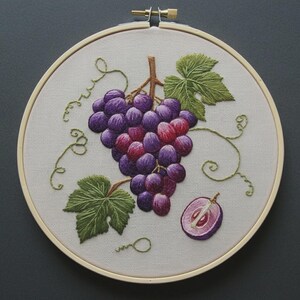 May include: A circular wooden embroidery hoop containing a fabric panel with a hand-stitched design of a cluster of purple grapes, two green leaves, and curling vines. A single halved grape is stitched to the lower right of the main cluster.