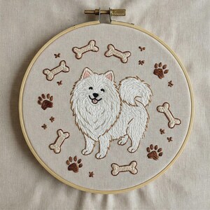Puede incluir: Un bastidor de bordado circular de madera que contiene un panel de tela con un diseño cosido a mano de un perro blanco esponjoso. El perro está rodeado de huellas de patas marrones y huesos de perro color crema sobre un fondo beige.