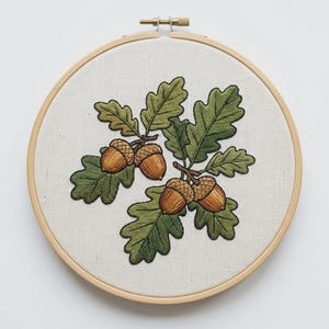 Può includere: Un telaio da ricamo circolare in legno contenente un tessuto color bianco sporco. Il tessuto presenta un dettagliato ricamo fatto a mano raffigurante quattro ghiande marroni attaccate a steli con diverse foglie di quercia verdi.