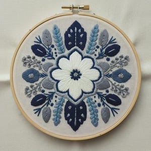 May include: A circular embroidery hoop containing a piece of off-white fabric with a symmetrical floral design. The pattern features a large central white flower surrounded by dark blue, light blue, and grey stitched leaves and small sprigs.