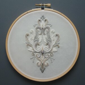May include: A circular wooden embroidery hoop containing white fabric with an intricate, symmetrical hand-stitched design in shades of silver, white, and cream thread, accented with small clear beads and a central faceted crystal embellishment.