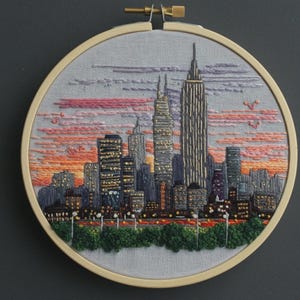 Könnte beinhalten: Ein runder Stickrahmen mit einer handgestickten Stadtsilhouette bei Sonnenuntergang. Die Szene zeigt hohe Wolkenkratzer in Grau- und Goldtönen vor einem Himmel aus rosa, orangefarbenen und violetten Fäden sowie grünes Blattwerk am unteren Rand.