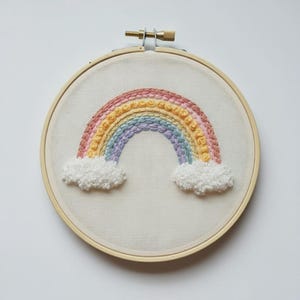 May include: A circular wooden embroidery hoop containing a piece of off-white fabric. The fabric features a hand-stitched rainbow design with textured, raised embroidery in shades of pink, yellow, blue, and purple, ending in white clouds.