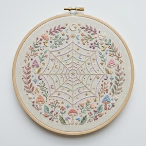 May include: A circular wooden embroidery hoop containing a piece of off-white fabric with a hand-stitched design. The center features a spider web surrounded by small stars and crescent moons, encircled by colorful mushrooms, leaves, and floral sprigs.