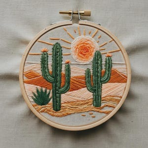 Op de afbeelding: Een ronde houten borduurring met een stoffen paneel waarop een geborduurd woestijnlandschap is afgebeeld. Het ontwerp bevat twee groene saguaro-cactussen, een kleine agaveplant en een feloranje zon met stralen boven zandduinen.