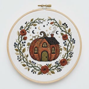 May include: An embroidery hoop containing white fabric with a hand-stitched design of a pumpkin-shaped house with a chimney, surrounded by a circular wreath of green vines, dark berries, and rust-colored roses, accented by small stars and moons.