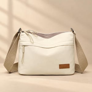 May include: A cream-colored crossbody bag featuring a wide, textured beige shoulder strap, multiple zippered compartments with matching pull tabs, and a small rectangular brown leather-like patch on the lower right corner.
