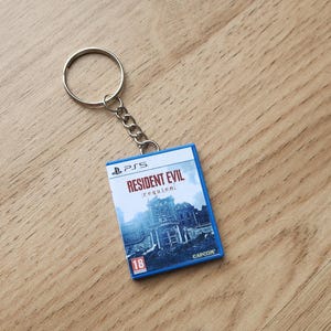 Op de afbeelding: Een miniatuur sleutelhanger in de vorm van een PlayStation 5-videogamehoesje voor Resident Evil Requiem. De rechthoekige hanger heeft een blauwe rand, het PS5-logo, de speltitel in rode letters, een gebouwillustratie en een 18-leeftijdslabel.