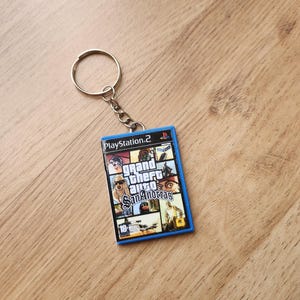 Può includere: Un portachiavi rettangolare che riproduce in miniatura la custodia del videogioco Grand Theft Auto San Andreas per PlayStation 2. Il ciondolo presenta un bordo in plastica blu e un anello in metallo con una corta catenella attaccata.