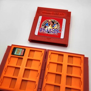 May include: A red Game Boy cartridge with a label featuring Charizard and the text Pocket Monsters, alongside an open orange storage case with multiple slots, one of which contains a small Animal Crossing Nintendo Switch game card.