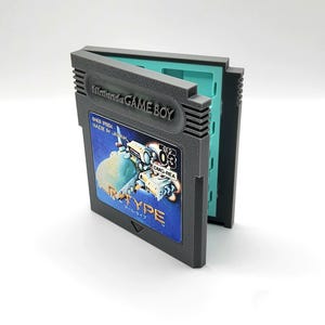 Può includere: Una custodia in plastica grigia per cartucce Nintendo Game Boy con design a cerniera. La parte anteriore presenta un'etichetta per il gioco R-Type, con il testo Nintendo GAME BOY, DMG-REA e 03. L'interno della custodia è di un colore verde acqua.
