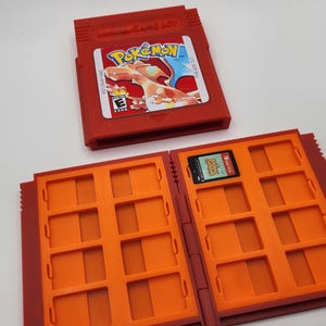May include: A red Nintendo Game Boy cartridge featuring a Pokémon label with Charizard and the ESRB E rating, positioned above an open red and orange plastic storage case with twelve slots, one of which contains an Animal Crossing game card.