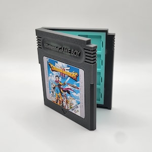 Può includere: Una custodia in plastica grigio scuro a forma di cartuccia per Nintendo Game Boy. La parte anteriore presenta un'etichetta per il gioco Dragon Warrior III con illustrazioni colorate dei personaggi e testo. La custodia si apre rivelando un interno azzurro.