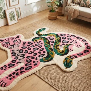 Puede incluir: Una alfombra con forma que presenta un leopardo rosa con manchas negras entrelazado con una serpiente verde y amarilla. La alfombra tiene un fondo blanco roto y sigue los contornos irregulares de las figuras animales como pieza decorativa.