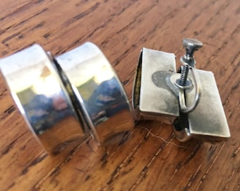 May include: A pair of silver clip-on earrings. The earrings are made of two stacked, rounded, silver pieces. The back of the earrings has a screw-on closure.