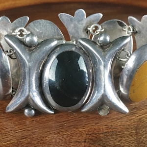 May include: A silver bracelet with three oval-shaped stones. The stones are red, black, and yellow. The bracelet has a decorative design with a floral motif.