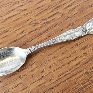 May include: A silver souvenir spoon with an image of the "Market Place Bldg." on the bowl and a depiction of a person on the handle.