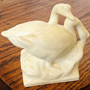 May include: A pair of white ceramic geese with their necks intertwined, standing on a base with a textured surface.