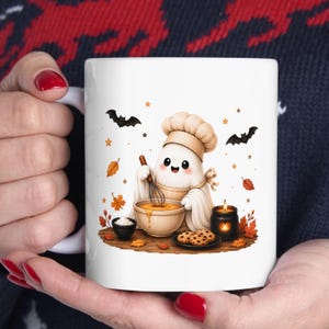 May include: A white ceramic mug featuring a cartoon ghost wearing a chef hat, whisking batter in a bowl. The design includes chocolate chip cookies, a small black candle, orange autumn leaves, stars, and two black bats on a wooden surface.