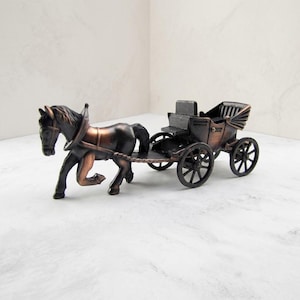 May include: A metallic bronze-toned figurine depicting a horse pulling a carriage. The horse is in a trotting pose, connected by a harness to a four-wheeled carriage with a high seat and a folding hood. The entire piece features a dark finish.