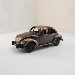 May include: A miniature model of a classic Volkswagen Beetle car featuring a weathered, metallic bronze and dark charcoal finish. The toy vehicle has rounded fenders, a curved roofline, and black wheels with a detailed tread pattern.
