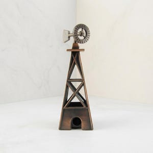 May include: A metallic windmill figurine with a bronze-toned finish, featuring a tall, open-frame tower structure with cross-bracing supports and a circular fan blade assembly at the top, resting on a base with a small, centered arched opening.