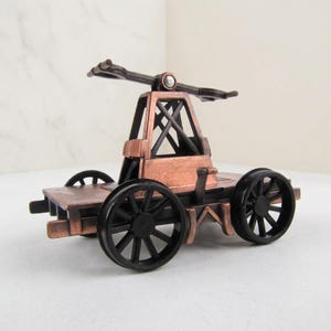 May include: A miniature metal handcar model featuring a copper-toned frame with black wheels and a central pumping handle mechanism. The structure includes a flat platform base and a vertical support frame with cross-bracing, finished in a metallic copper.