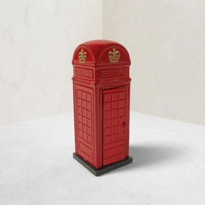 May include: A bright red miniature telephone box figurine featuring a domed roof, a rectangular door with a grid pattern, and gold-colored crown emblems embossed above the word TELEPHONE on the front panel, all set on a dark gray base.
