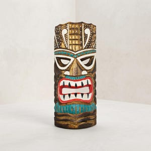 May include: A hand-carved wooden tiki mask featuring a dark brown finish with painted accents in white, turquoise, and red. The mask displays a stylized face with large white eyes, a wide mouth with sharp teeth, and intricate geometric patterns.