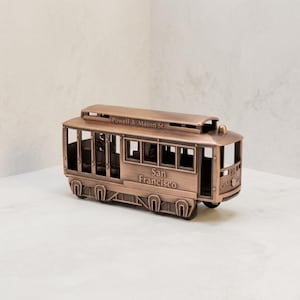 May include: A copper-toned metal model of a San Francisco cable car. The side features the text Powell & Mason St. and San Francisco, while the front displays the number 88 and the words Cable Car in raised lettering on the metallic surface.