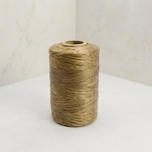 May include: A cylindrical spool of light brown raffia twine. The twine is tightly wound around a central core, creating a textured, fibrous surface with a consistent tan color throughout the entire length of the spool of craft material.