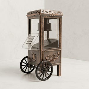 May include: A bronze-colored popcorn machine with a vintage cart design, featuring black wheels, clear glass panels, and the word Popcorn embossed on the side. The unit has a hinged clear plastic door and a dark interior compartment for popping corn.