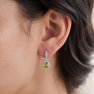 May include: A silver drop earring featuring a square-cut, light green peridot gemstone at the centre. The stone is surrounded by a halo of small, clear, sparkling stones, with additional clear stones set along the vertical silver bail.