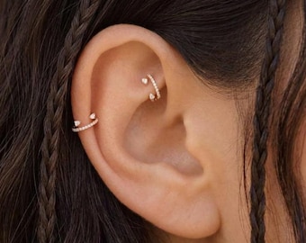 May include: Two gold-toned ear cuffs featuring small, clear, sparkling stones. One cuff is positioned on the rook, while the other is placed on the helix, both showcasing a delicate, curved design that hugs the cartilage of the ear.