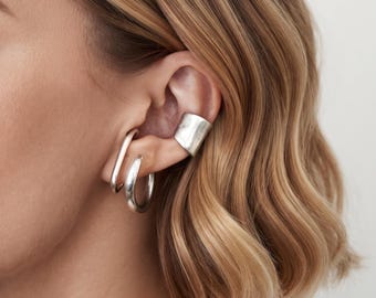 May include: A set of three silver-toned earrings consisting of a smooth, wide ear cuff positioned on the upper cartilage, a sleek oval-shaped hoop, and a thick, rounded circular hoop earring worn on the lower earlobe for a layered appearance.