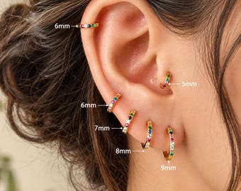 May include: A set of gold hoop earrings featuring multi-colored gemstones. The hoops are labeled with sizes 5mm, 6mm, 7mm, 8mm, and 9mm. Each earring is adorned with small, vibrant stones in various colors set along the circular gold band.