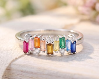 May include: A silver-toned ring featuring a row of multi-colored baguette-cut gemstones in shades of red, pink, orange, yellow, green, and blue, accented with small round clear stones set between the colored gems.