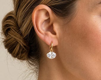 May include: A gold hoop earring featuring a dangling oval-shaped clear gemstone. The earring is crafted from polished yellow metal and showcases a single faceted stone that reflects light, suspended from a simple circular hoop closure.