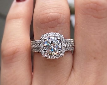 May include: A silver-toned ring featuring a large, round, brilliant-cut center stone surrounded by a halo of smaller round stones. The band is composed of three distinct rows, each encrusted with tiny, sparkling pavé-set stones.