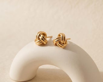 May include: A pair of gold stud earrings featuring a polished knot design. Each earring consists of a small, rounded metallic knot shape with a post backing, showcasing a smooth, reflective surface and a warm, consistent yellow gold tone throughout.